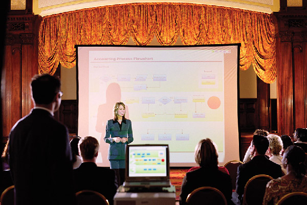 Presenter standing in front of a projected flowchart, speaking to a seated audience in a ballroom.